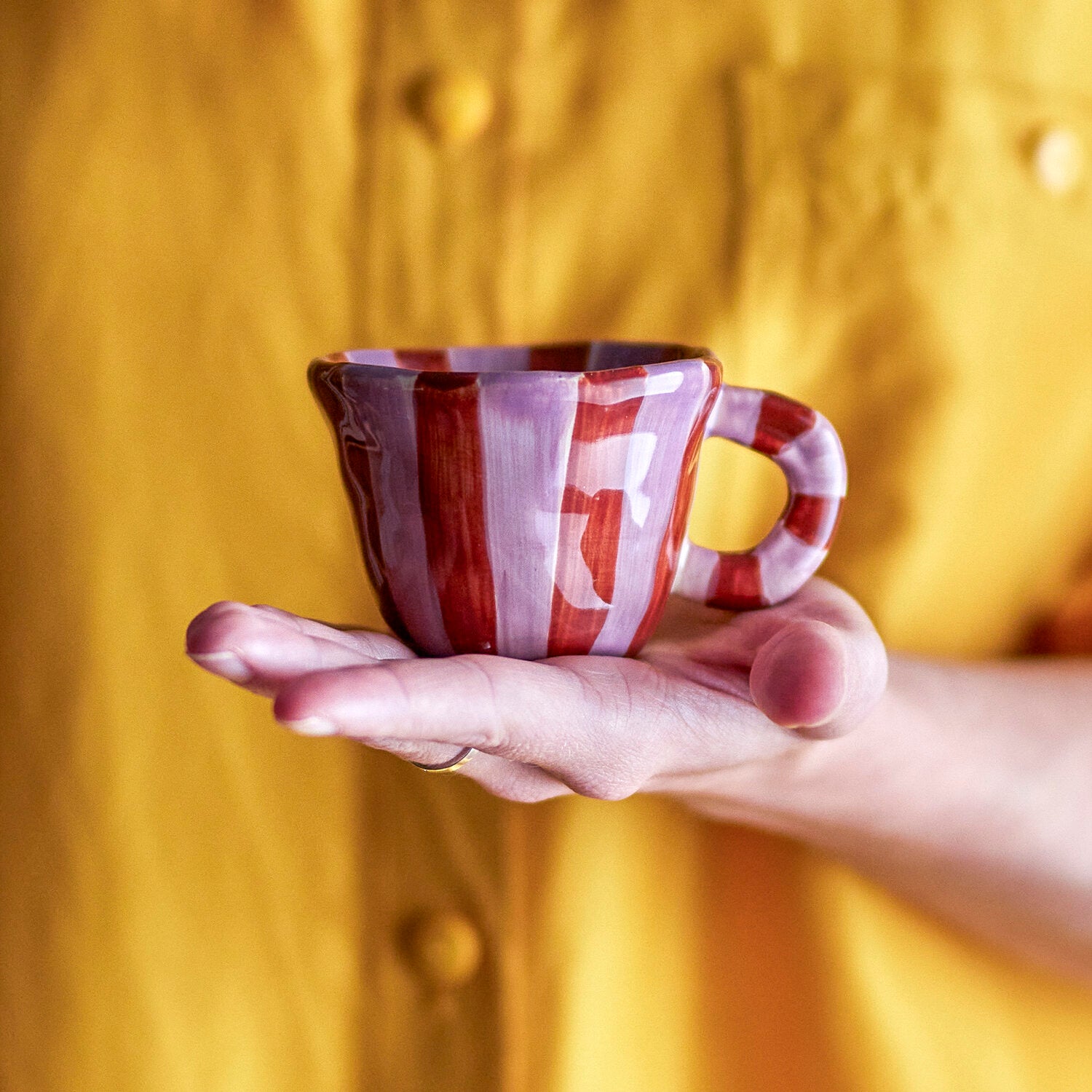 Tasse aus Keramik mit rot-violetten Vertikalstreifen und abgesetztem Henkel, in Hand