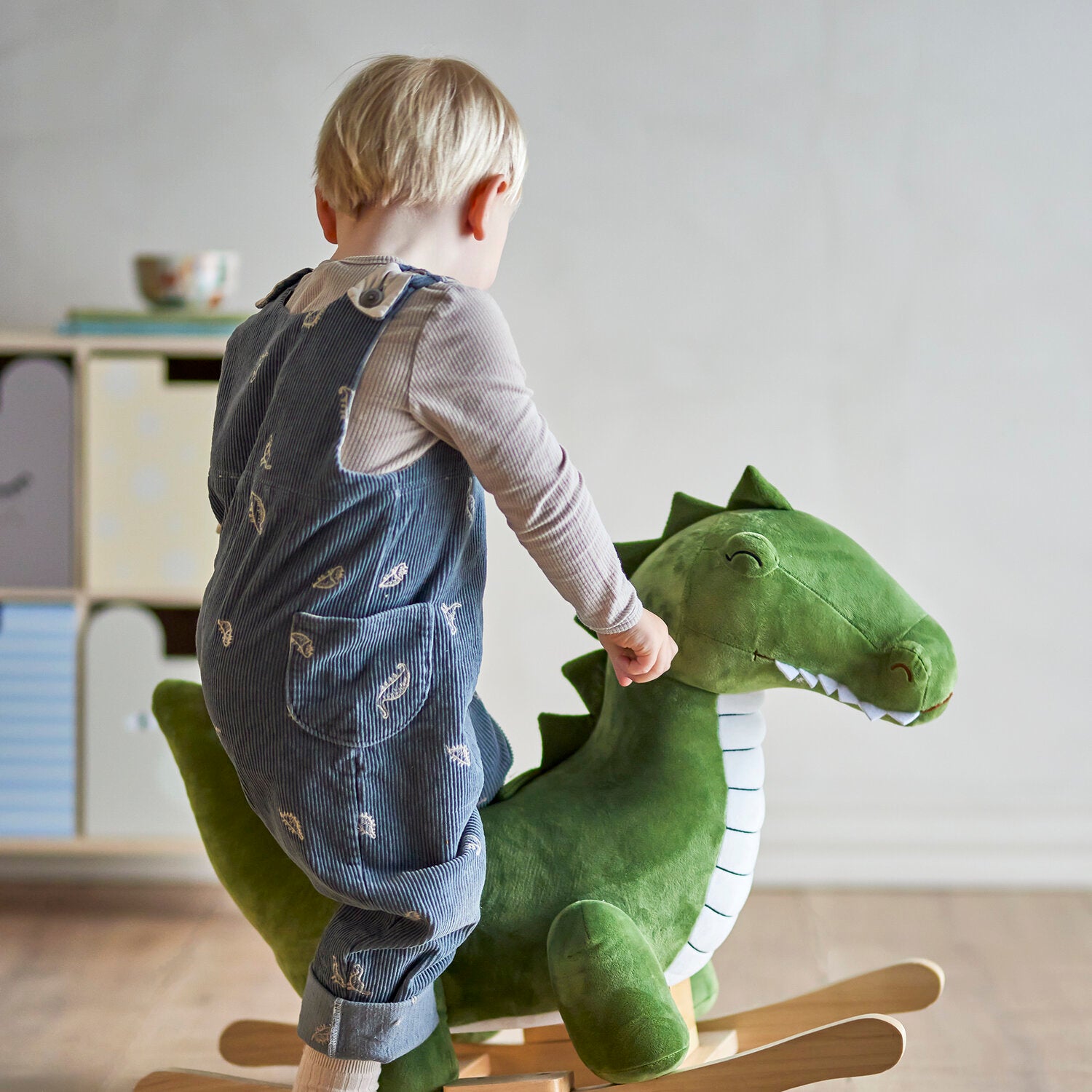 Kind mit Jeans-Latzhose schaukelt grünes Plüsch-Krokodil auf Holzschaukel