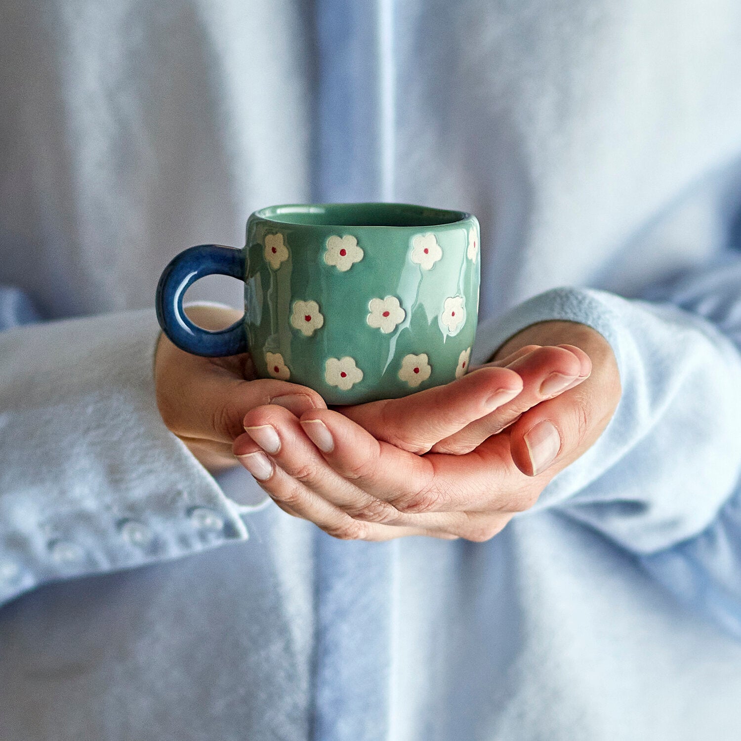 Grüne Keramiktasse mit Henkel und weißen Blüten, von Händen gehalten