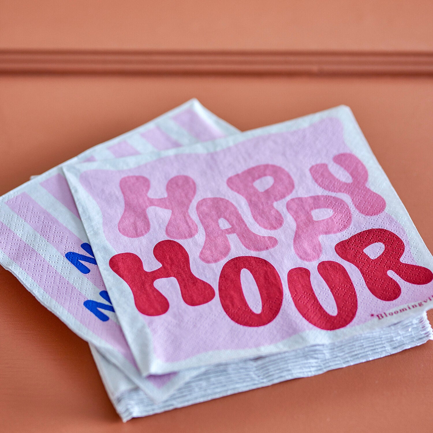 Gestapelte Papierservietten, lilafarbener Hintergrund mit „HAPPY HOUR“-Schrift in Pink