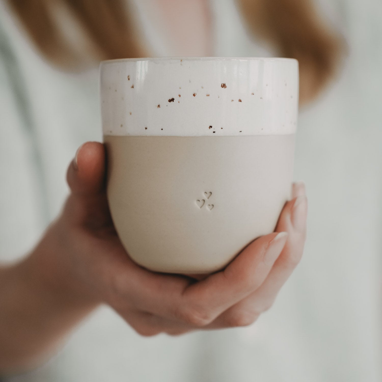 Kleiner Becher in Naturfarbe, von beiden Händen gehalten; weißes Band mit Herz-Reliefs.