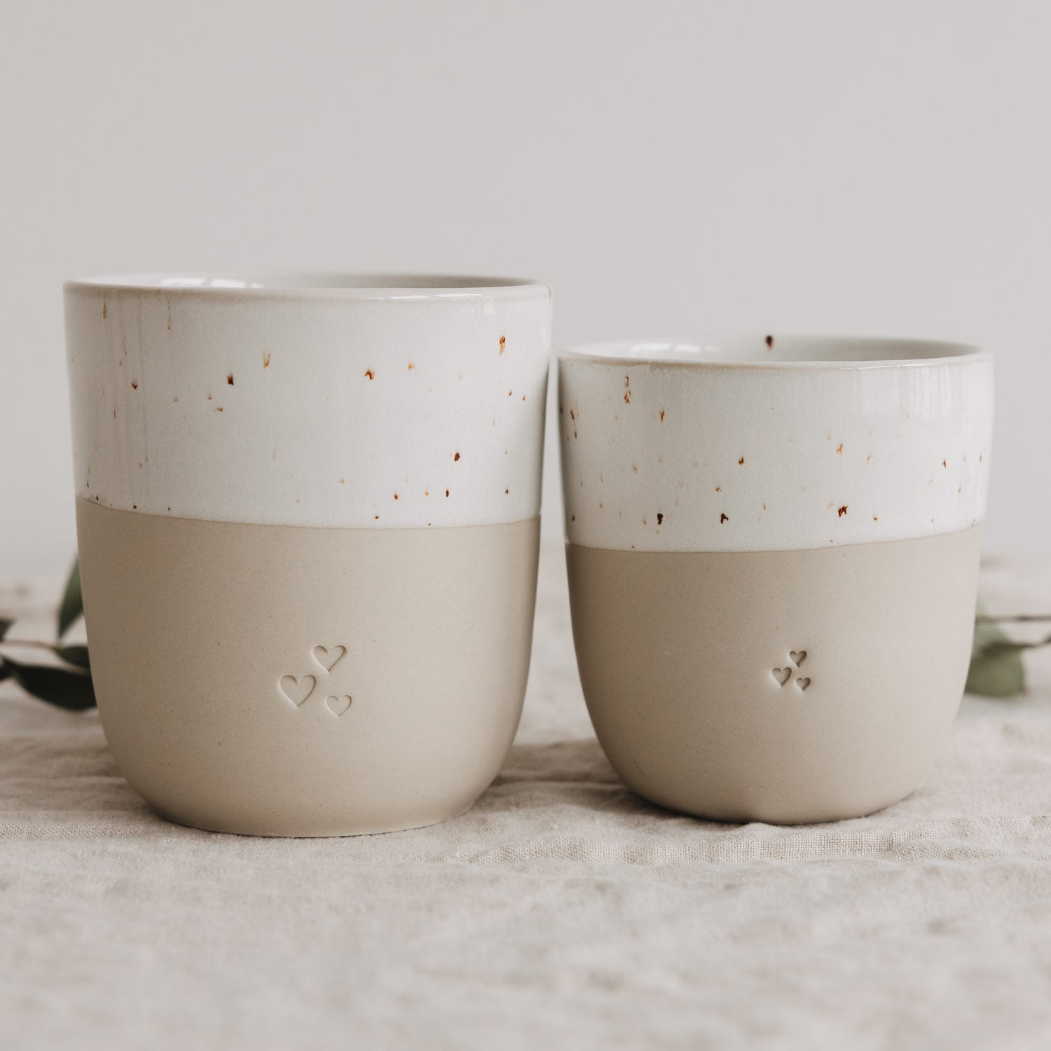 Zwei kleine Becher in Naturfarbe mit weißem Band und Herz-Reliefs, nebeneinander.