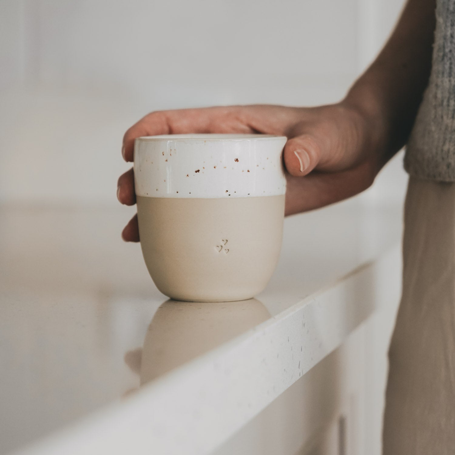 Kleiner Becher in Naturfarbe, von einer Hand gehalten; weißes Band mit Herz-Reliefs.