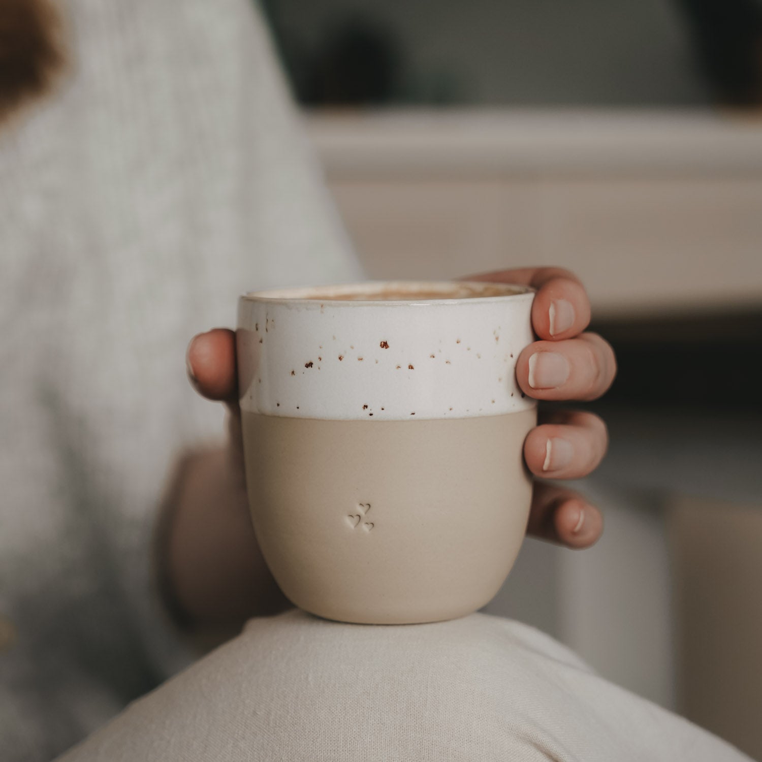 Kleiner Becher in Naturfarbe, von einer Hand gehalten; weißes Band und Herz-Reliefs.