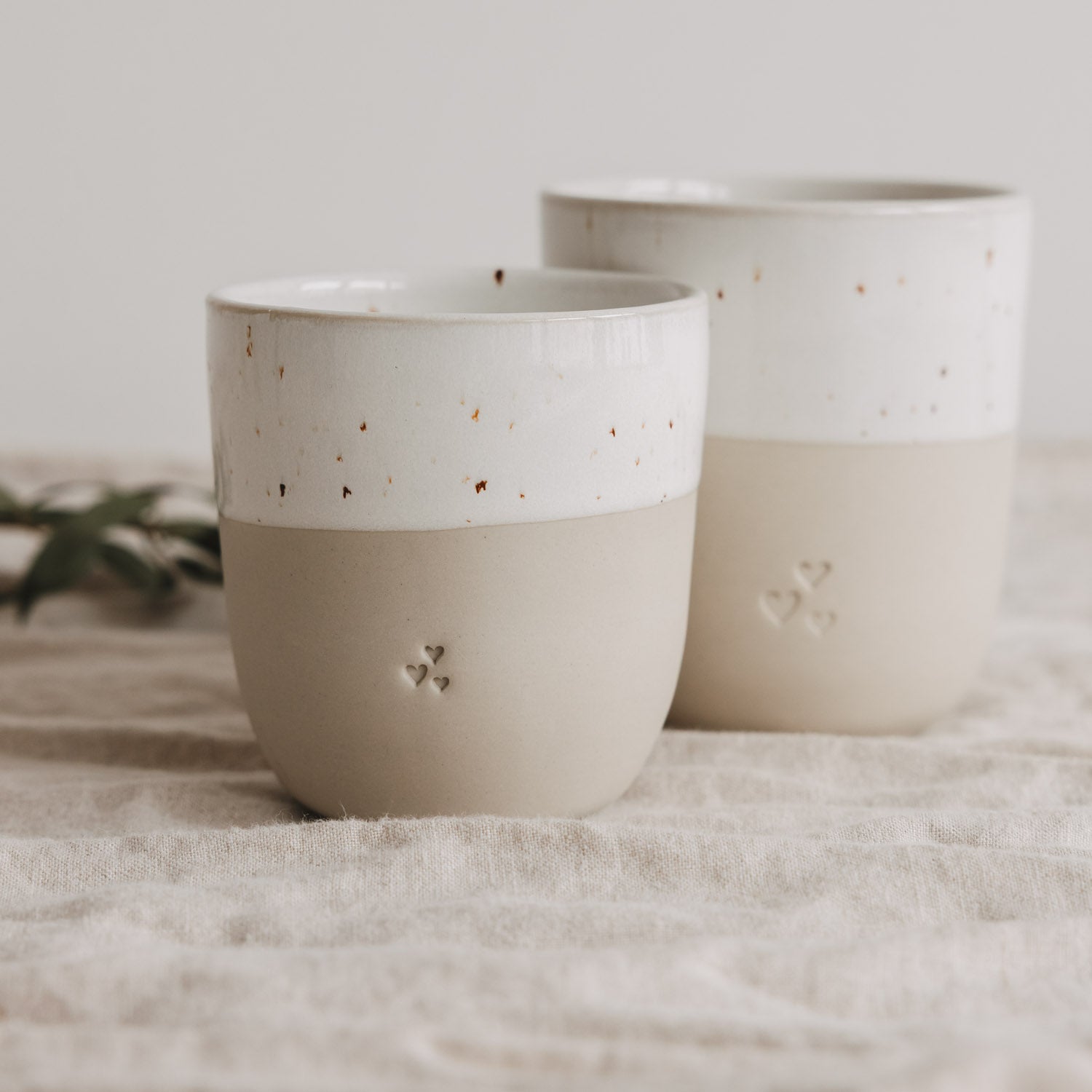 Zwei kleine Becher in Naturfarbe mit weißem Band und Herz-Reliefs, schräg zueinander.
