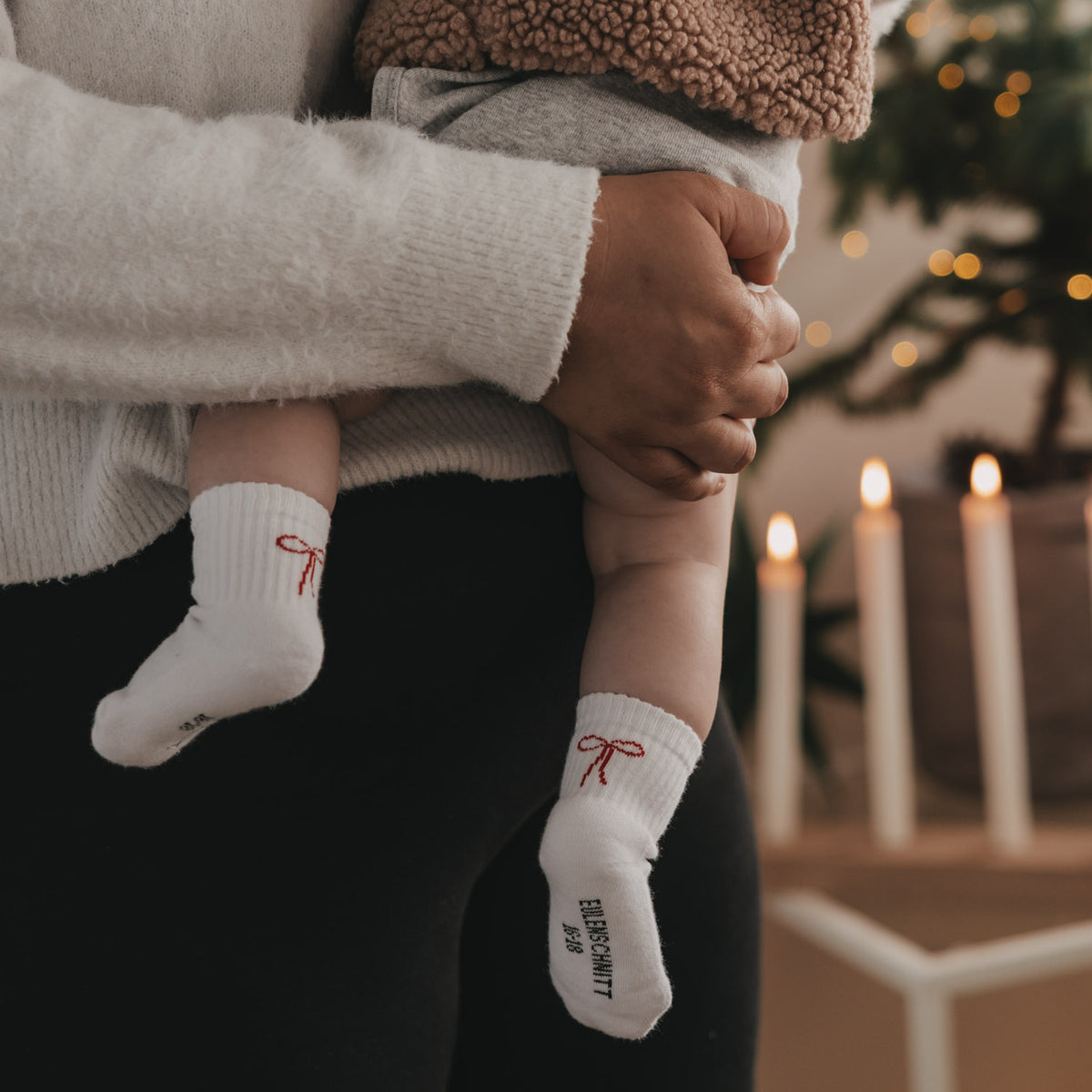 Babysocken in Weiß mit roter Schleife und Schriftzug, seitliche Ansicht am Knöchel