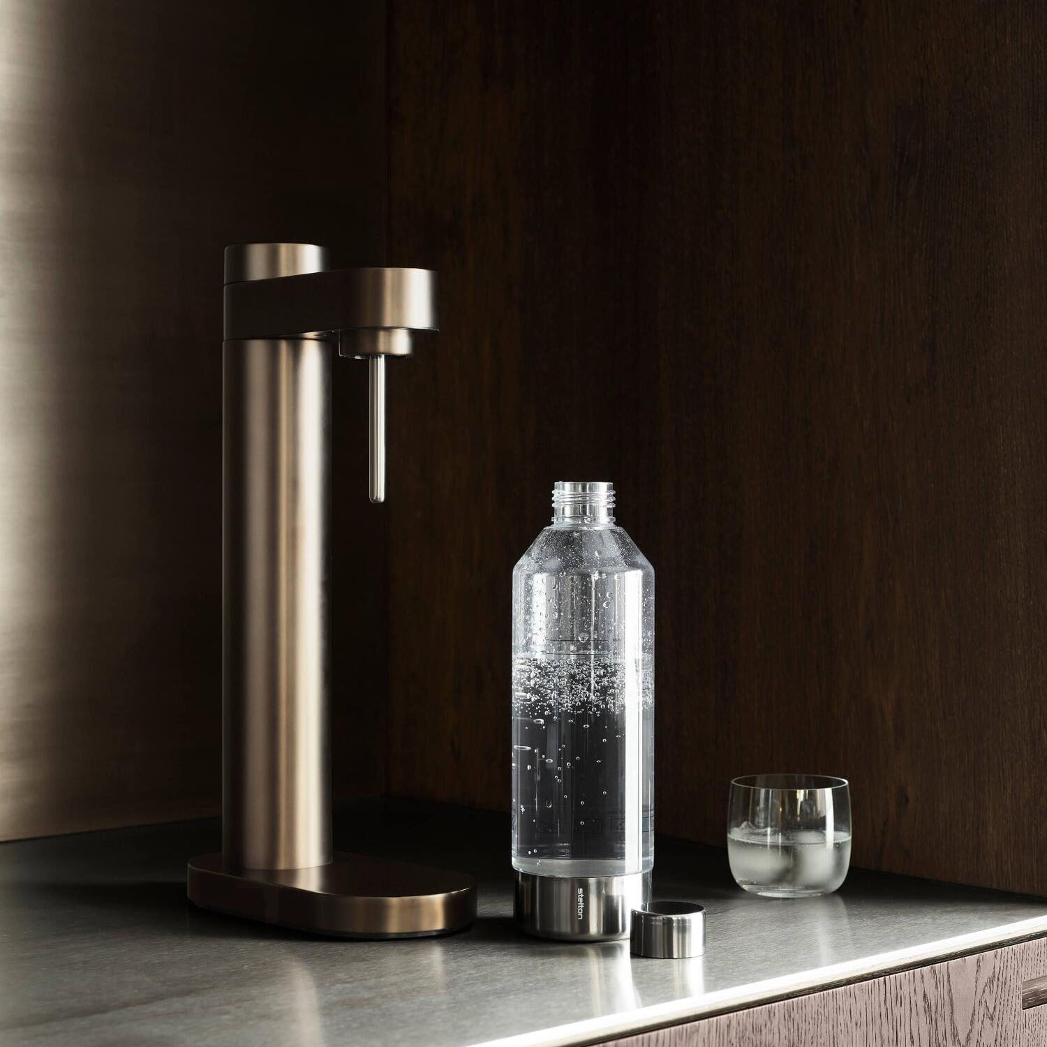 Wassersprudler auf Sockel mit mattbraunem Metallstand, Glasflasche und Auslaufrohr