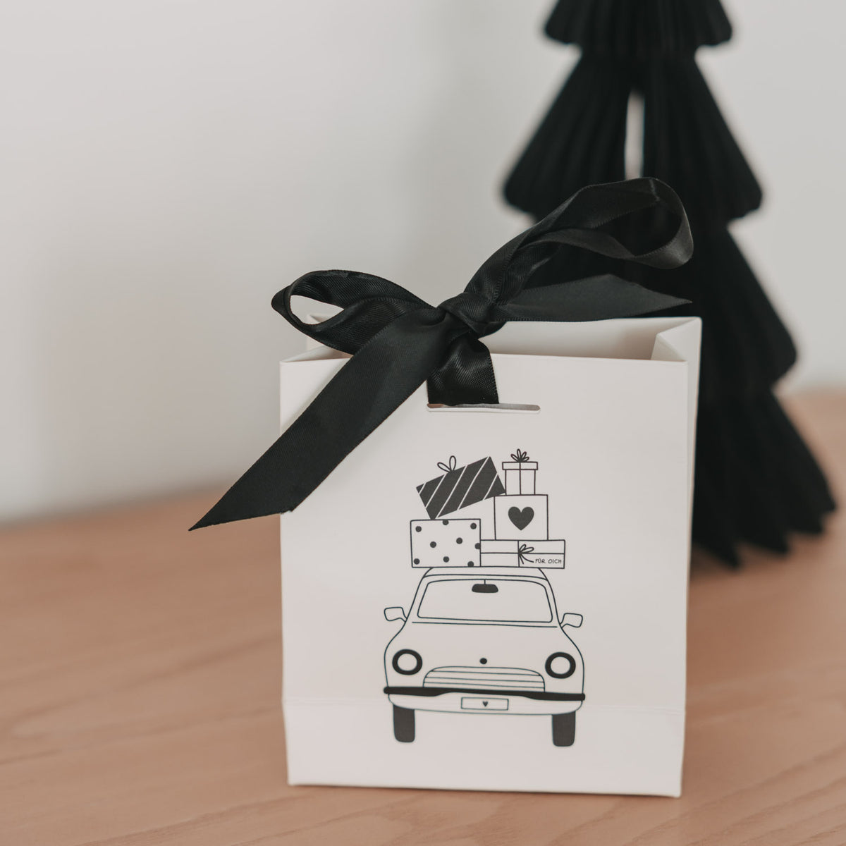 Weiße Mini-Geschenktüte mit schwarzer Schleife und Weihnachtsauto-Motiv auf braunem Boden