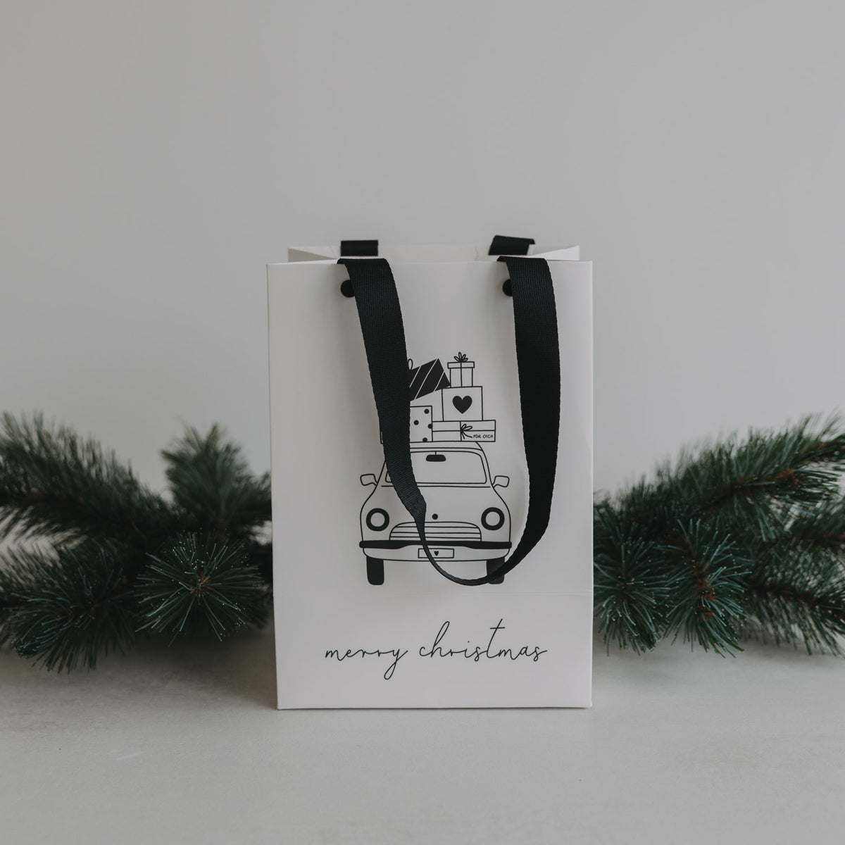 Weiße Geschenktüte mit schwarzem Trageband und Weihnachtsauto-Print; „merry christmas“ unten.