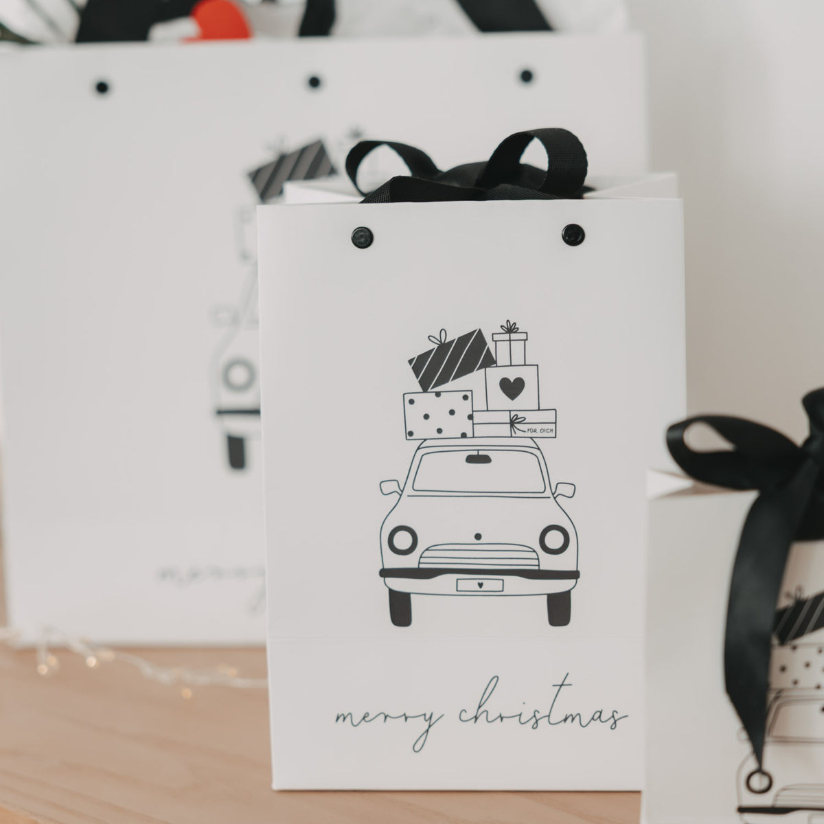 Weiße Geschenktüten im Set, Weihnachtsauto-Motiv mit schwarzem Trageband und „merry christmas“.