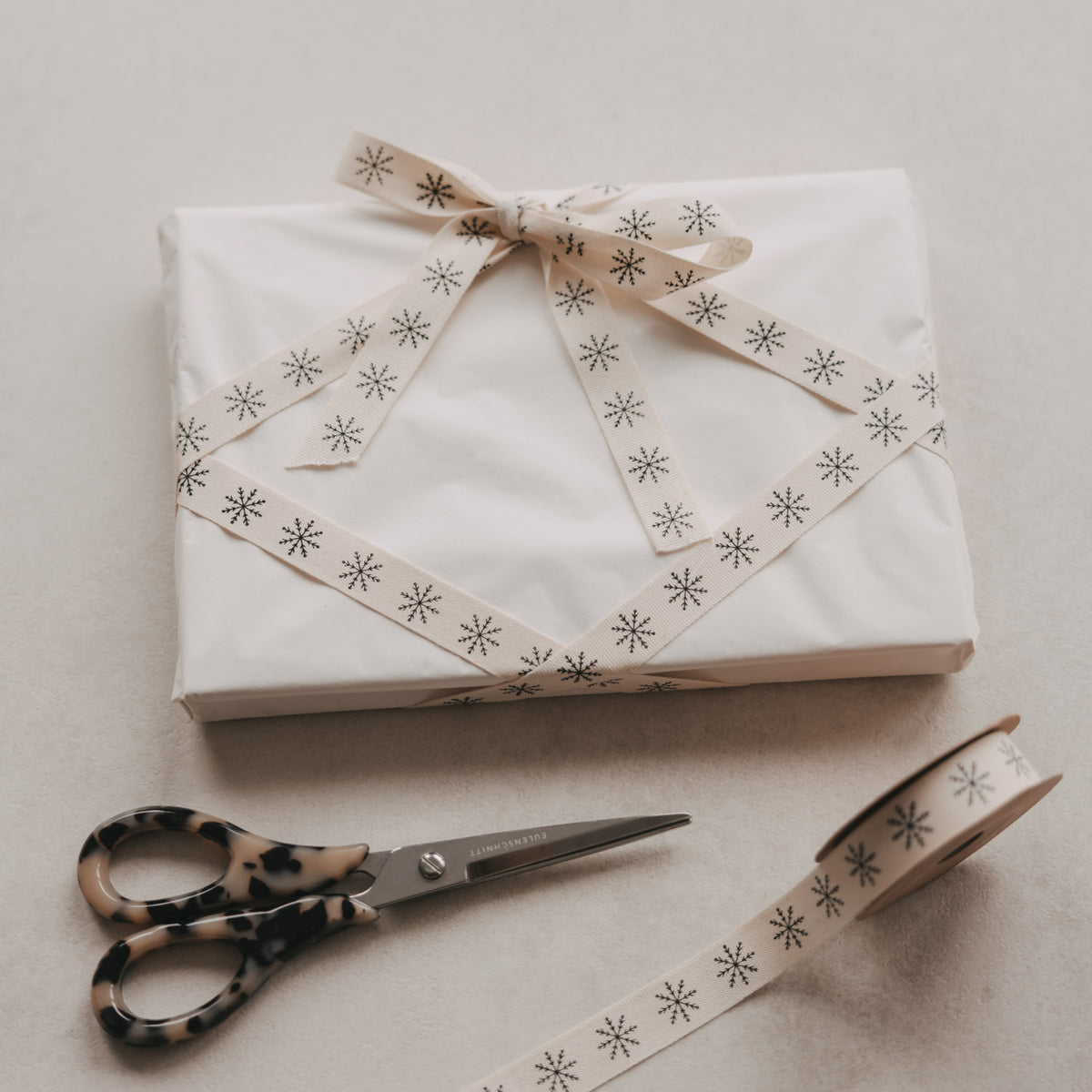 Geschenkband mit Schleife, beiger Stoff mit Schneeflocken, daneben Schere und Band